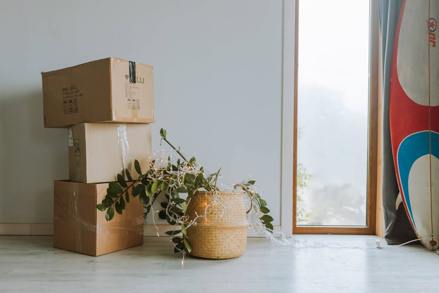 Une scène intérieure cosy avec des boîtes en carton, une plante et une planche de surf près d'une fenêtre.